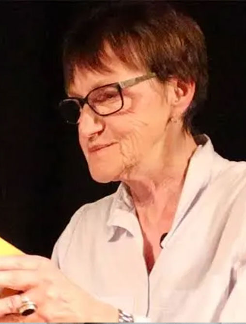 Elfriede Jelinek liest aus einem gelben Buch, sie trägt eine weiße Bluse und eine Brille.